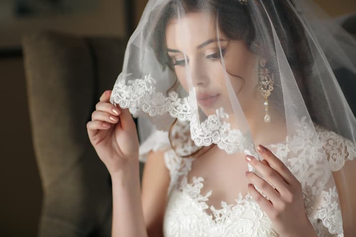 The young beautiful woman, the brunette with a wedding hairdress and a professional make-up, in ears expensive earrings, is dressed in a white wedding dress and a transparent white veil, waits for the groom in a hotel room