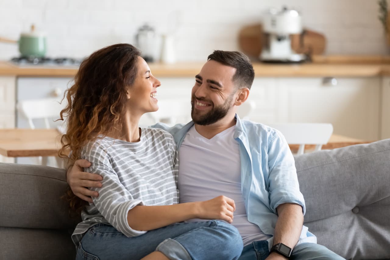 Happy young couple of man and woman embracing look at each other sitting on couch in living room. Attractive smiling husband and wife laughing having fun free time together.