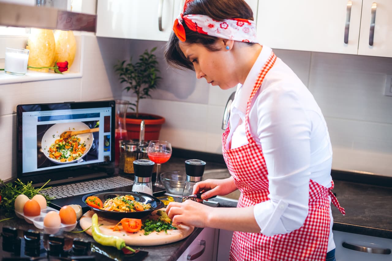 Young cute caucasian woman watching tutorial and making a meal in kitchen.
