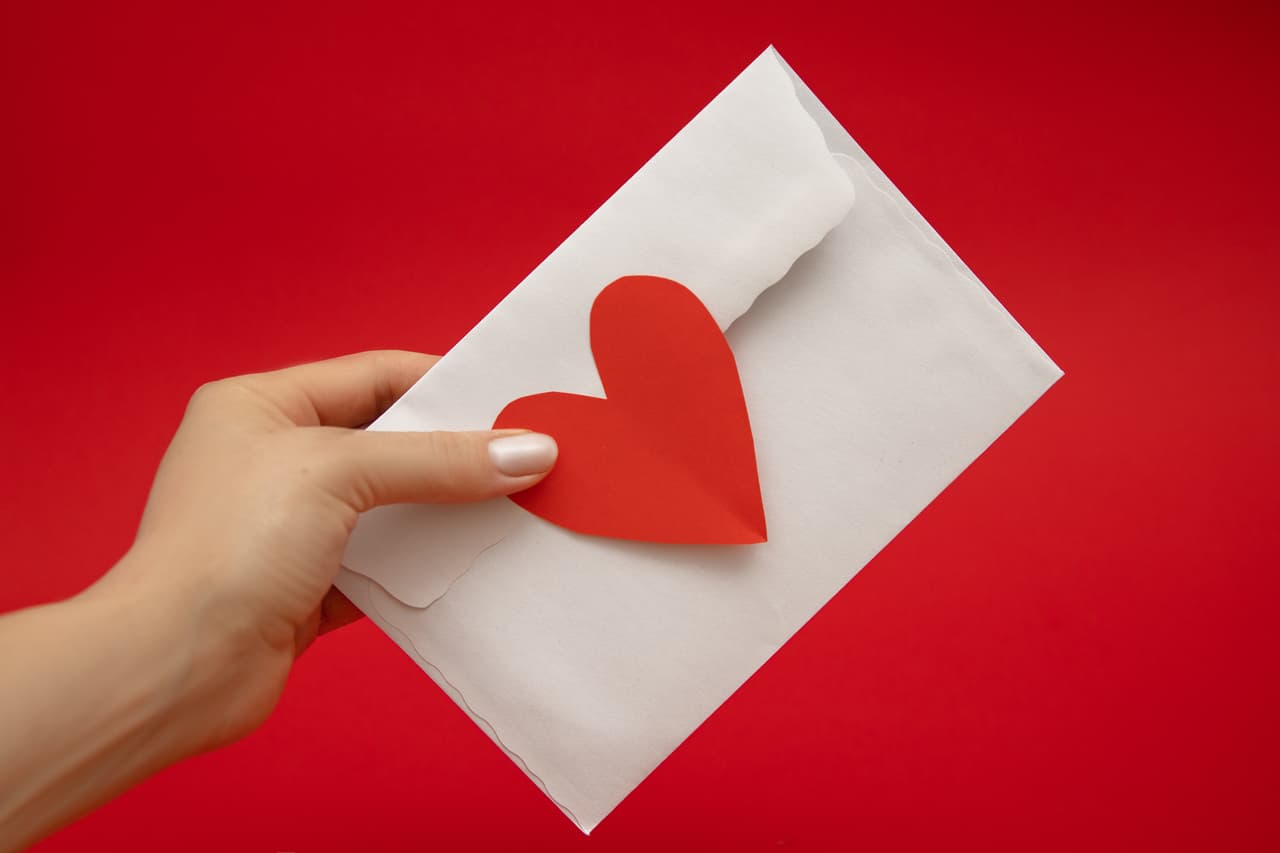 Woman's Hand holding envelope sealed with love in shape of red valentine's heart on red background. Valentine's Day
