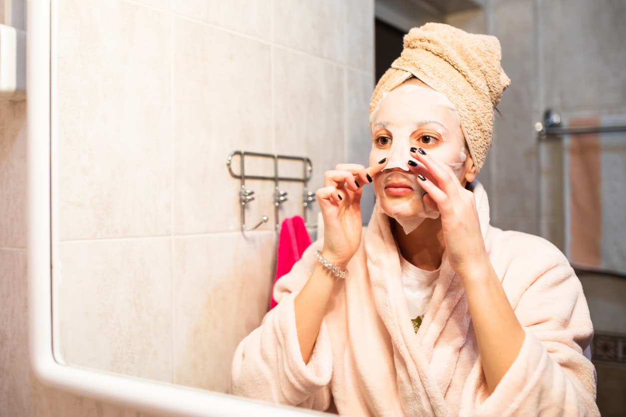 A beautiful young woman is applying a sheet mask in front of the mirror