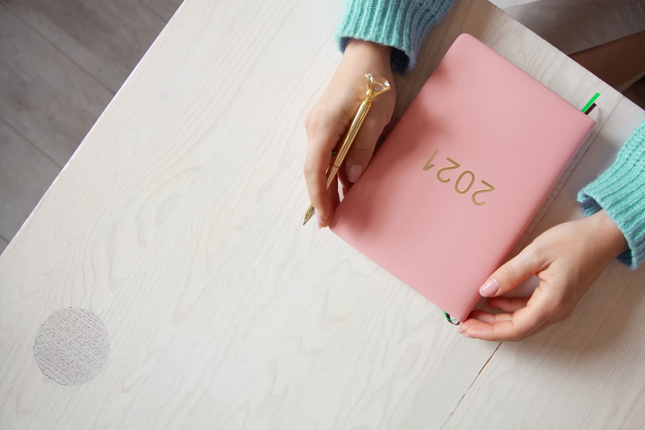 Top view of woman hands in warm sweater with coral coloured 2021 diary book on table. Future plans and achievements for new year 2021. Wellbeing lifestyle