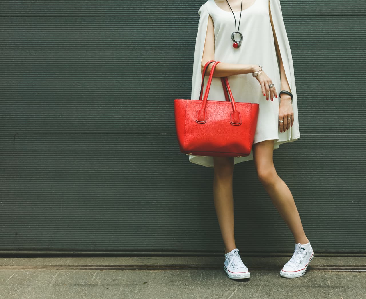 Fashionable beautiful big red handbag on the arm of the girl in a fashionable white dress and sneakers, posing near the wall on a warm summer night. Warm color.