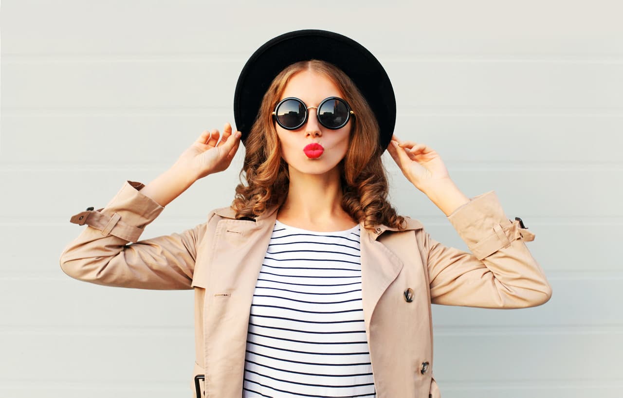 Fashion portrait pretty sweet young woman blowing red lips wearing a black hat sunglasses coat over grey background