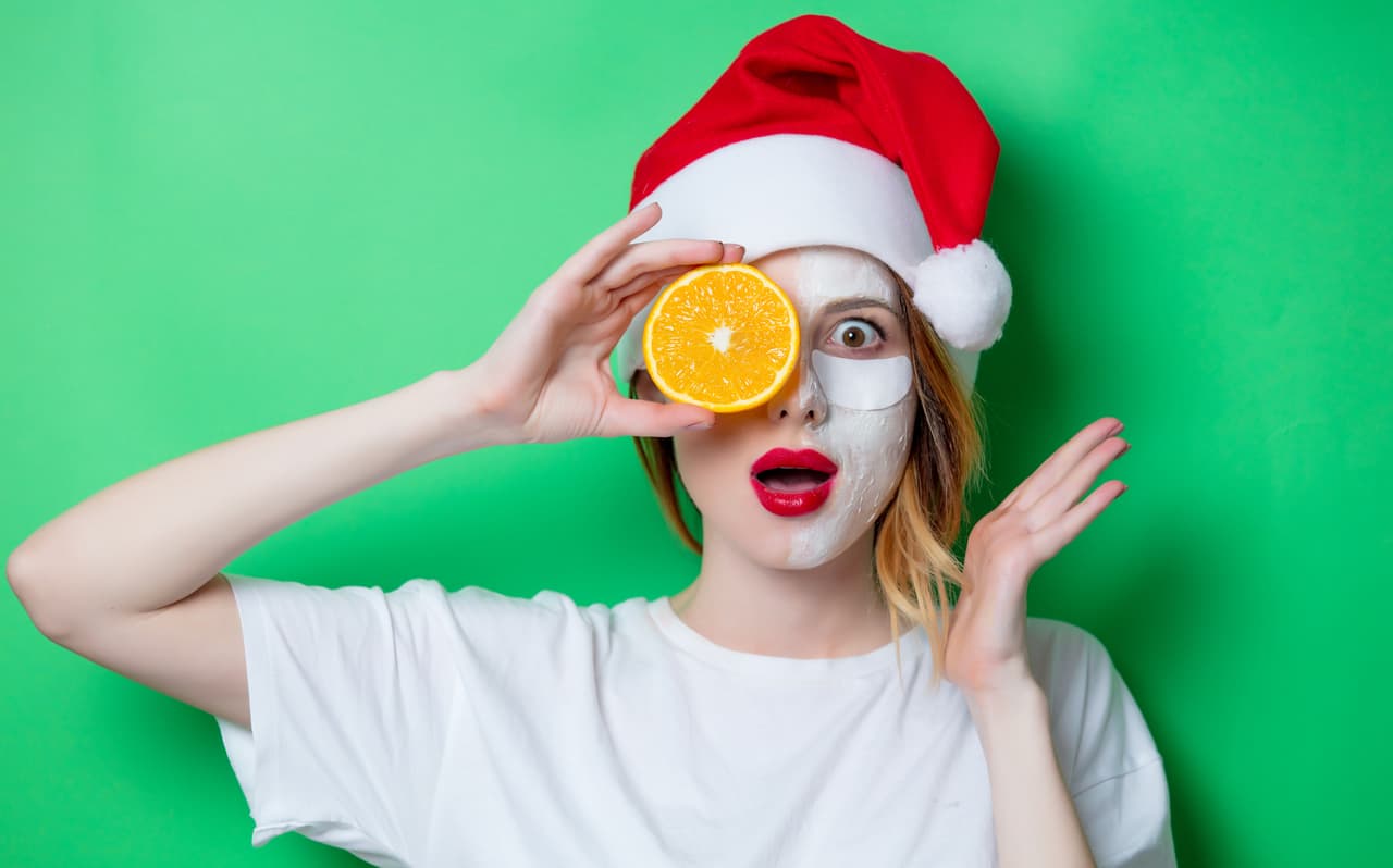 Woman using eye patch for her eyes in Santa Claus hat with cream on her face and holding orange. Portrait of natural and true female as usually they are looks like