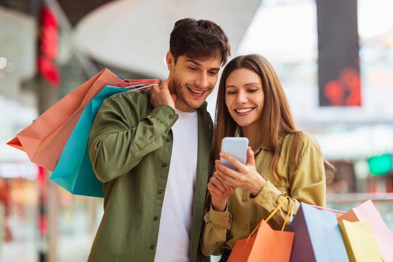 Happy Couple Shopping Using Mobile Phone Application Holding Colorful Shopper Bags And Buying Clothes In Modern Mall On Weekend. Customers Hold Smartphone Browsing Internet For Sales. Ecommerce