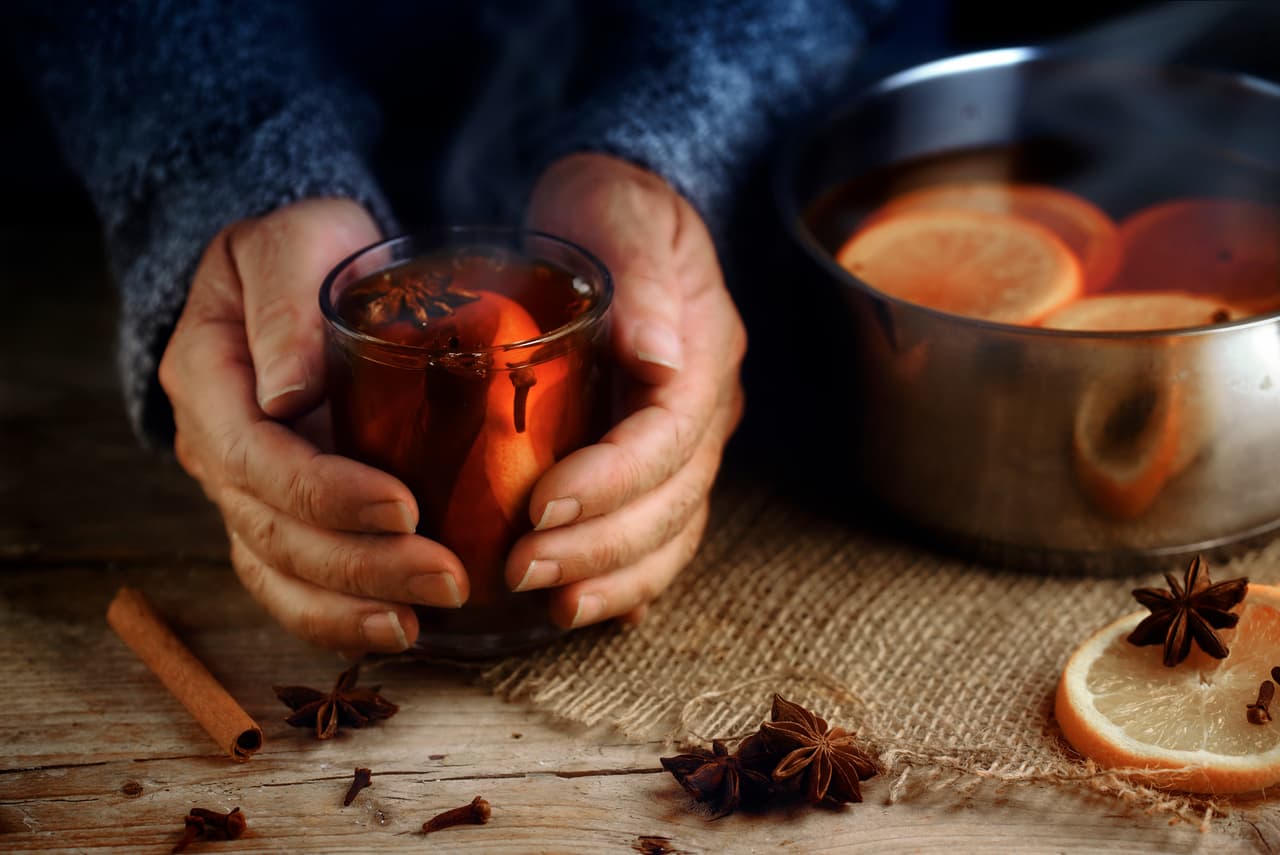 Older female hands holding a glass mug with hot mulled wine next to the steaming cooking pot, Christmas ingredients, orange slices, cinnamon sticks, star anise and cloves, a warming home scene on a rustic wooden table, selected focus, narrow depth of field