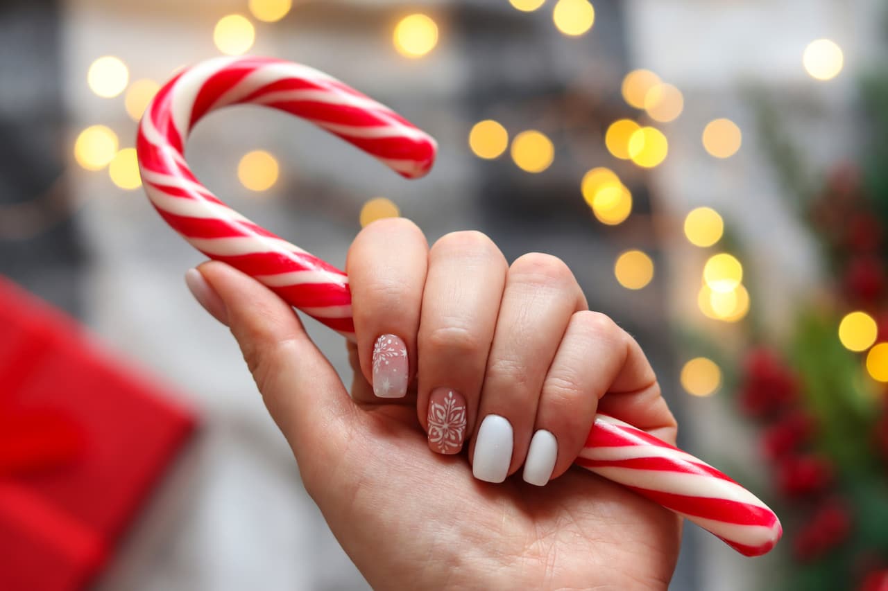 Woman's hand with gel polish manicure white color and with snowflakes ornament, holding candy cane against festive Christmas background. Selective focus. Idea of the winter manicure.