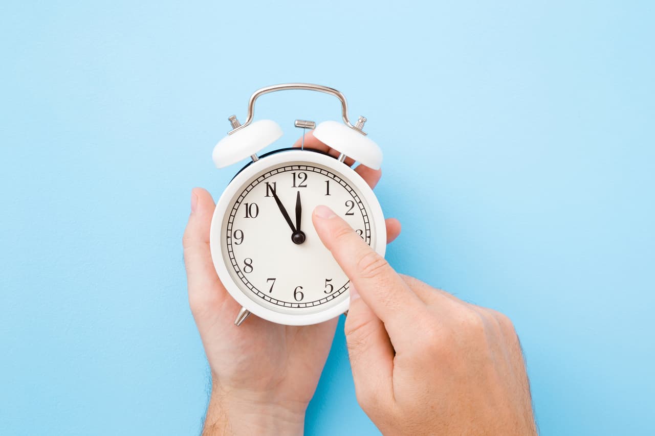 Man hand holding white alarm clock. Finger pointing to arrow of twelve o'clock. Light pastel blue background. Time change concept. Closeup. Top view.