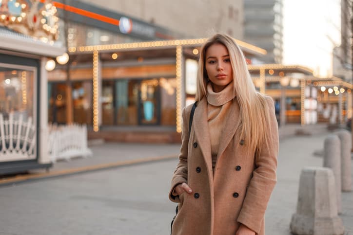 Charming young beautiful woman in a stylish coat and a vintage knitted sweater walking near the shops of the festive on the street. Urban fashion elegant girl blonde. Have a good day