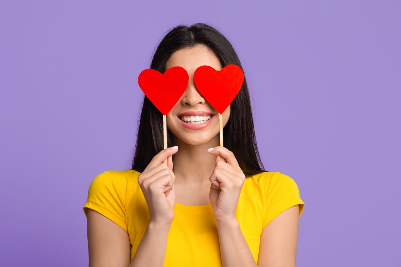 All You Need Is Love. Romantic asian woman posing with red hearts over eyes and smiling, having fun, celebrating valentine's day alone, standing over purple studio background, closeup shot, free space