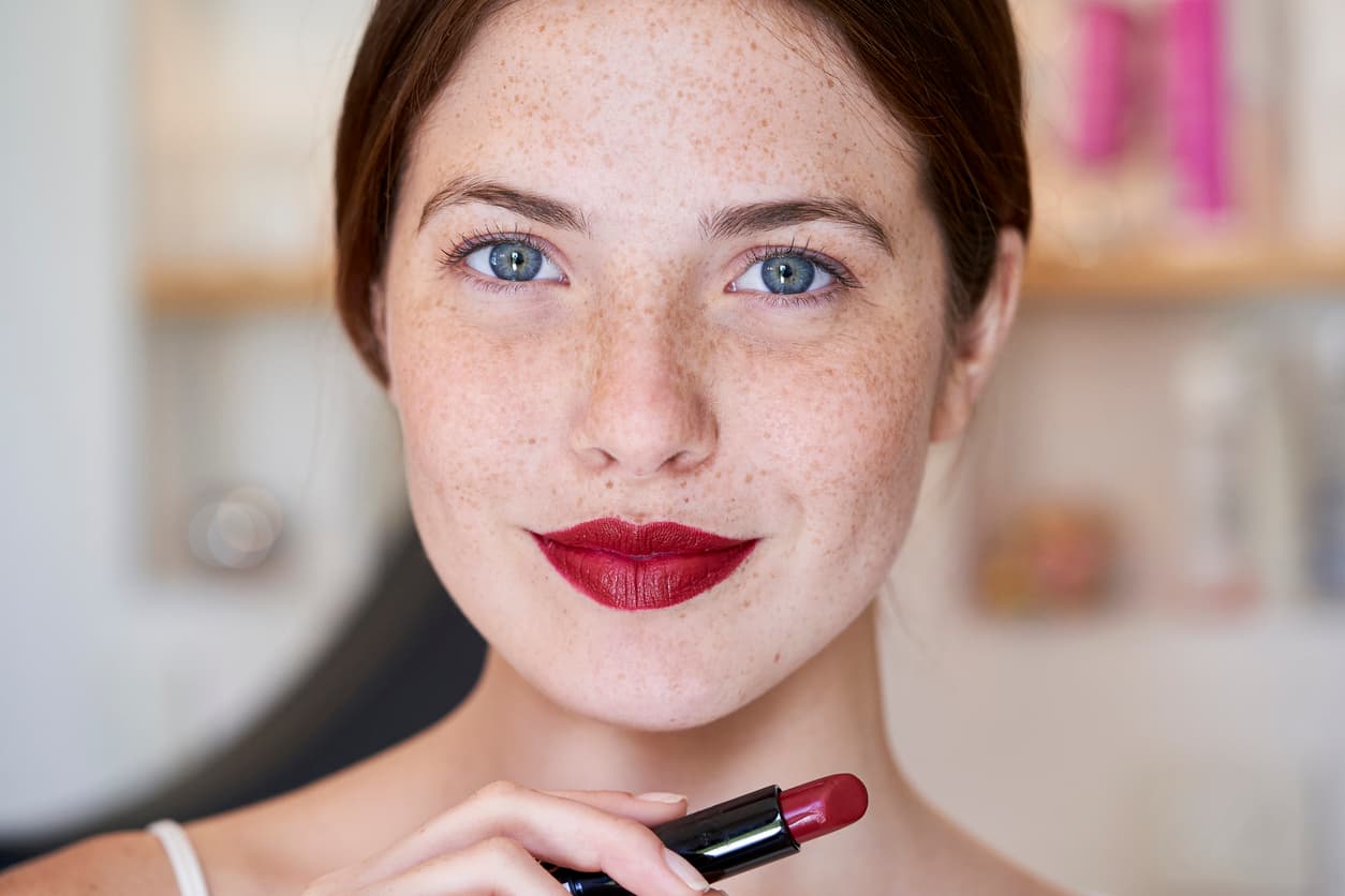 Portrait of smiling young woman with freckles holding maroon lipstick in hand while standing in bathroom