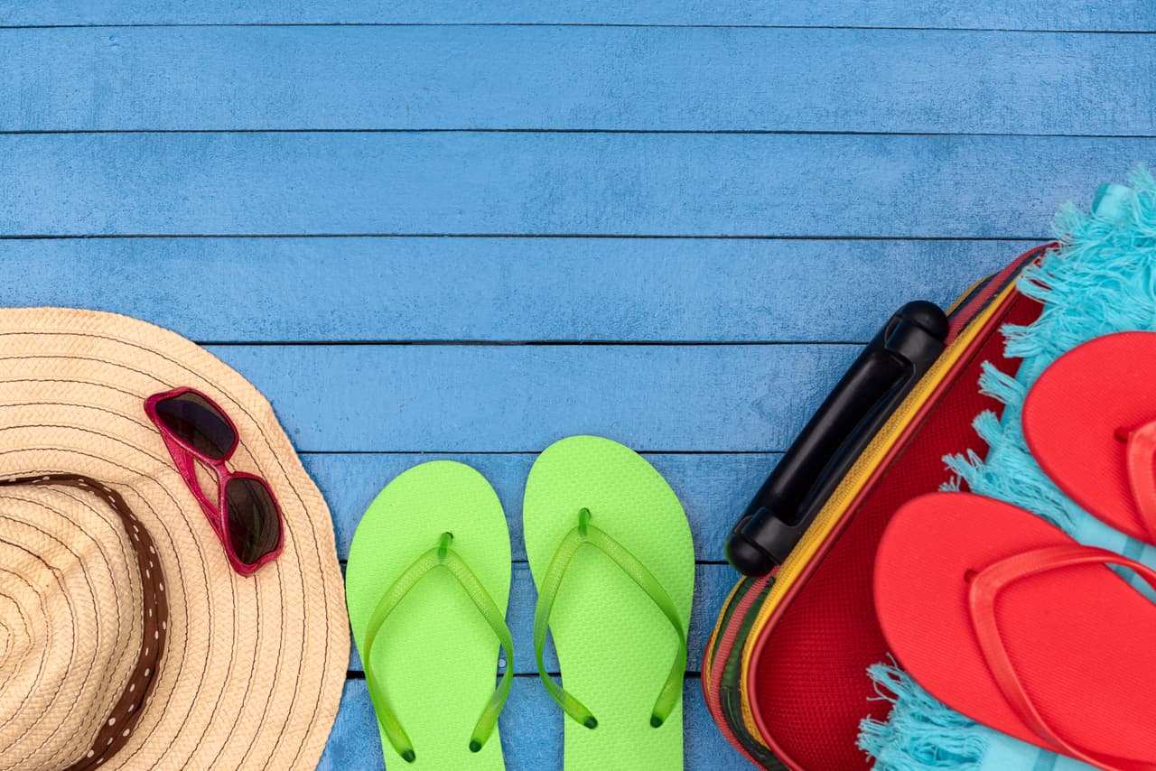 Suitcase with flip-flops, hat and sunglasses on wooden table.