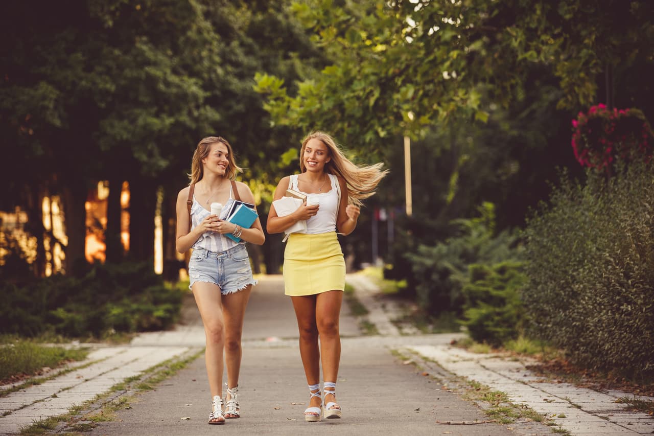 Front view of two smiling young women walking in campus, carrying books and backpacks.