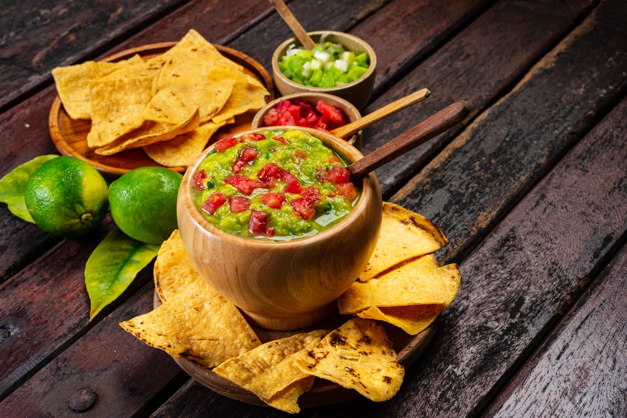 Typical mexican food of guacamole with tortilla chips ,cilantro , tomato, lemon and chives over rustic wooden table in the kitchen.This guacamole the ingredients are avocado, salt, juice of lemon, onion and tomato.