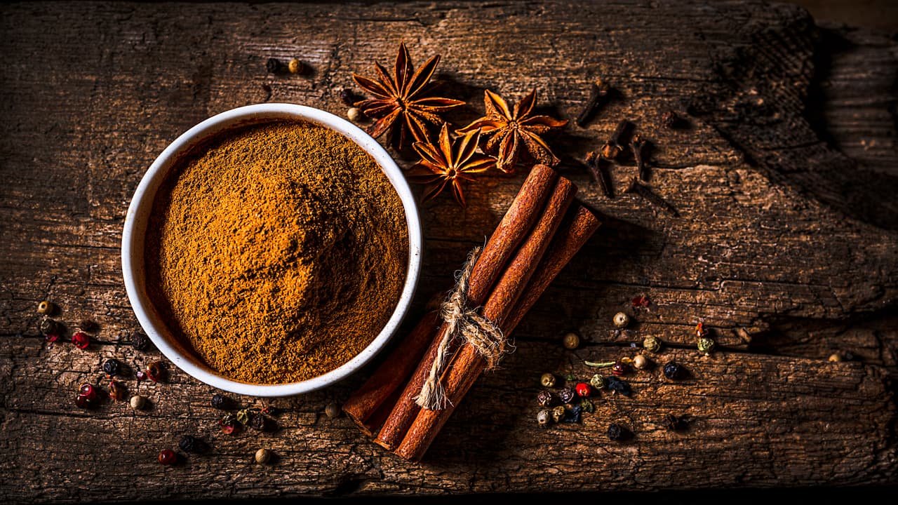 Scented spices background: top view of a white bowl filled with cinnamon powder shot on rustic wooden table. Cinnamon sticks, star anise, cloves and peppercorns are all around the bowl. Predominant color is brown. The composition is at the left of an horizontal frame leaving useful copy space for text and/or logo at the right. High resolution 16:9 format studio digital capture taken with SONY A7rII and Zeiss Batis 40mm F2.0 CF lens