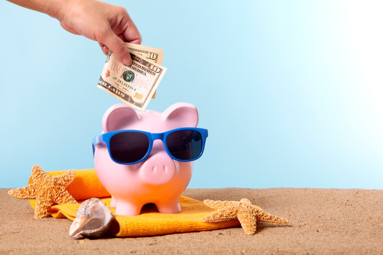 Saving for beach vacation, with pink piggy bank and sunglasses. Studio shot with plain blue background. Sharp focus on the ten dollar bill. Space for copy. Warm color and directional lighting are intentional.