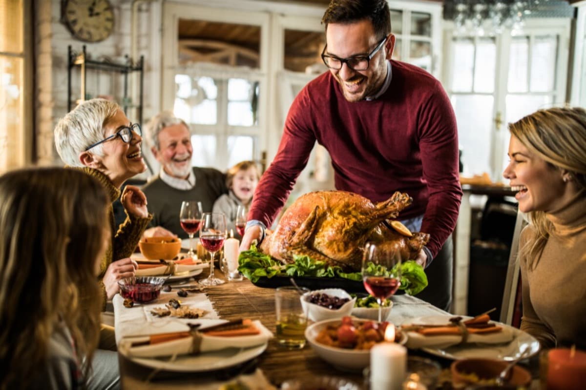 Happy man serving roasted turkey to his family during Thanksgiving dinner at dining table.
